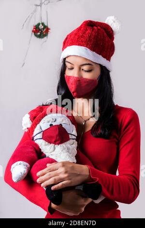 Jeune fille latine en tenue de Noël portant un Père Noël fabriqué, la fille et le Père Noël portent tous deux des masques chirurgicaux rouges Banque D'Images