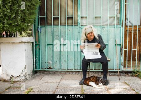 Une femme âgée lit le journal à sa porte tandis que son chat dort à ses pieds à Mtskheta, Géorgie, Caucase, Europe de l'est. Banque D'Images