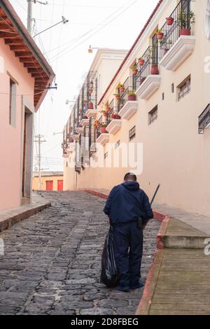 Homme faisant le nettoyage de la rue pavée de la ville coloniale - homme ramassant les ordures avec un sac et un balai dedans rue du centre historique de Quetzaltenango Banque D'Images