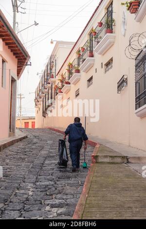 Homme faisant le nettoyage de la rue pavée de la ville coloniale - homme ramassant les ordures avec un sac et un balai dedans rue du centre historique de Quetzaltenango Banque D'Images