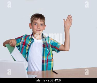 Un garçon élégant et mignon a soulevé sa main, tient dans son autre main un ordinateur portable avec des devoirs terminés, un ordinateur portable sur la table Banque D'Images