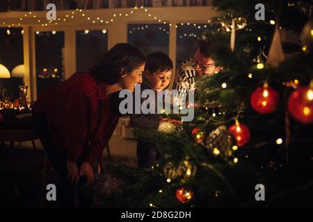 Un garçon regardant les lumières sur l'arbre de Noël avec ses parents debout. Photo intérieure d'une maison familiale décorée pour Noël. Banque D'Images