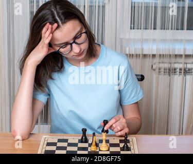 Caucasienne fille avec des lunettes pensant jouer aux échecs, jouant aux échecs à la maison pendant la quarantaine