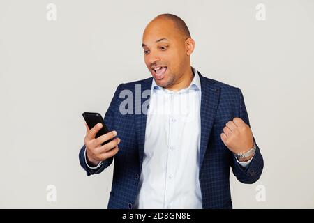 Portrait d'un jeune homme d'affaires afro-américain heureux utilisant un smartphone, criant avec fierté et célébrant la victoire et le succès, très excité, se réjouissant des émotions. Debout sur un fond gris. Banque D'Images