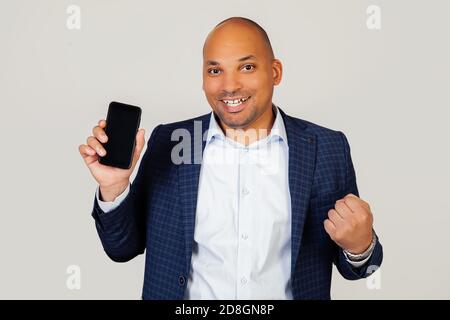 Portrait du jeune homme d'affaires afro-américain heureux, montre l'écran de smartphone, crie avec fierté et célèbre la victoire et le succès, très excité, se réjouit des émotions. Debout sur un fond gris Banque D'Images