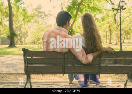 Vue arrière de multi ethnic couple assis sur banc en bois dans l'amour tout en appréciant la vue panoramique sur le parc verdoyant paisible Banque D'Images