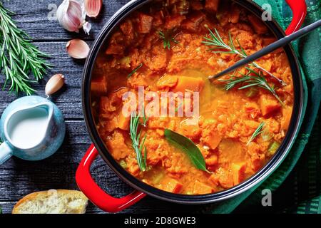 Soupe de potiron saine et à base de keto dans une casserole rouge avec une cuillère à verser sur un fond en bois avec pain grillé, crème, ail, feuille de Laurier, romarin, dessus Banque D'Images