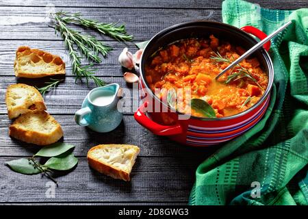 Soupe de potiron saine et à base de keto dans une casserole rouge avec une cuillère à verser sur un fond en bois avec pain grillé, crème, ail, feuille de Laurier, romarin, dessus Banque D'Images