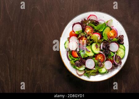 Salade végétale saine de tomates cerises, tranches de concombre, feuilles de laitue verte et violette, oignons et huile d'olive dans l'assiette sur table en bois vue du dessus Banque D'Images