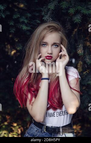 Portrait rapproché de la jeune fille blonde avec les yeux bleus avec mains près du visage à l'ombre des plantes vertes Banque D'Images