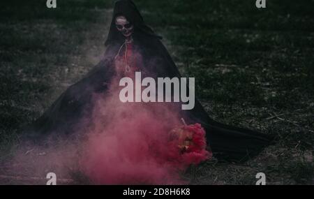 La jeune femme est assise parmi les champs dans le costume d'Halloween de la mort près de allongé sur la Jack-o-lanterne au sol avec de la fumée rouge. Concept Halloween. Crâne de sucre Banque D'Images