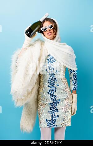 femme mécontentée dans des lunettes de soleil tenant une bouteille de champagne sur le bleu Banque D'Images