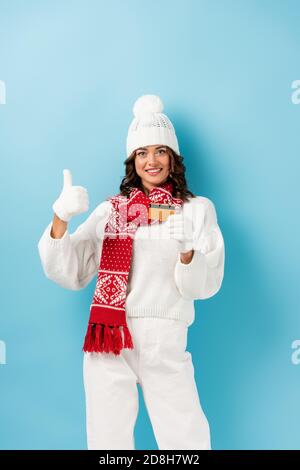 bonne jeune femme dans la tenue d'hiver tenant une carte de crédit et avec le pouce vers le haut en bleu Banque D'Images