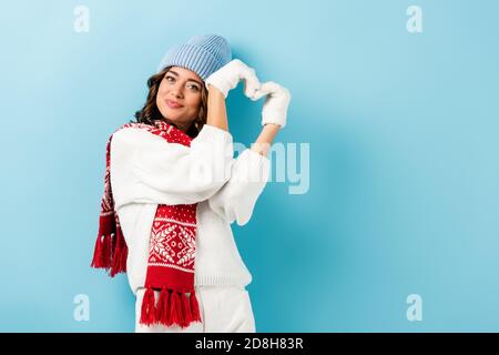 jeune femme heureuse en tenue d'hiver, écharpe chaude, gants et chapeau montrant le coeur signe sur bleu Banque D'Images