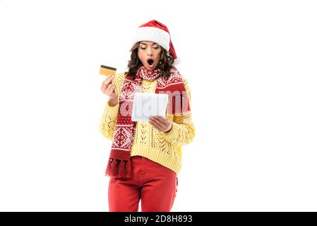 une jeune femme choquée regarde le chapeau et le foulard de santa tablette numérique tout en tenant la carte de crédit isolée sur blanc Banque D'Images