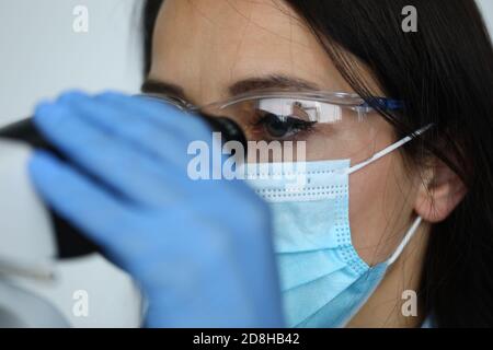 Femme en lunettes de sécurité et masque médical sur le visage au microscope dans le concept de laboratoire biologique Banque D'Images