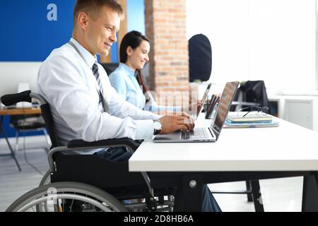 Un jeune homme souriant en fauteuil roulant travaille sur un ordinateur portable au bureau avec une femme collègue Banque D'Images