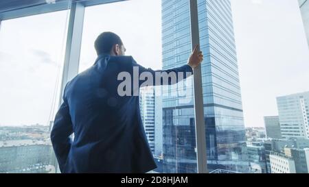 Homme d'affaires sérieux, vêtu d'un costume debout dans son bureau, regardant par la fenêtre et envisageant le prochain contrat Big Business. Ville principale Banque D'Images