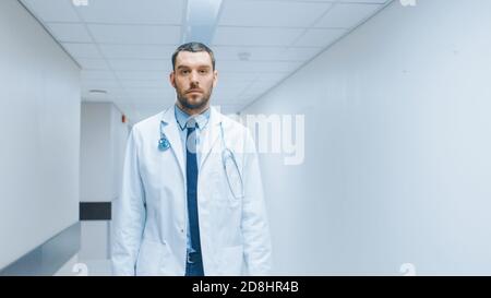 Un beau médecin portant un manteau blanc avec stéthoscope se promène dans le couloir de l'hôpital. Clinique moderne et lumineuse avec un personnel professionnel. Banque D'Images