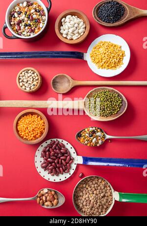 Légumineuses variété de légumineuses sur fond de table rouge. Pelles, bols et louches avec des aliments sains végétariens secs. Vue de dessus, plan d'appartement Banque D'Images