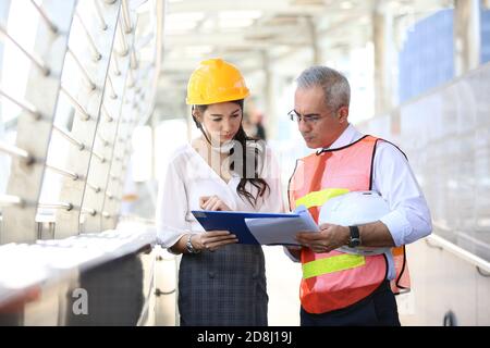 Femme ingénieur industriel portant un casque blanc lorsqu'elle se trouve sur un chantier de construction, avec un homme d'affaires qui parle du plan de travail, de l'ingénieur et de l'archite Banque D'Images
