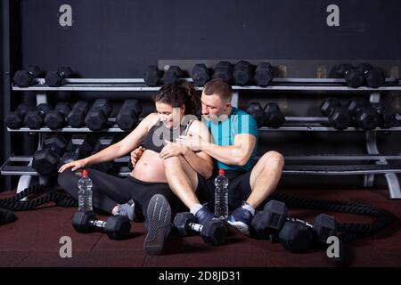 Une femme enceinte va faire du sport avec son mari en salle de sport pour un entraînement multidisciplinaire. Couple athlètes s'attendant à bébé tout en se reposant après l'entraînement et les exercices Banque D'Images