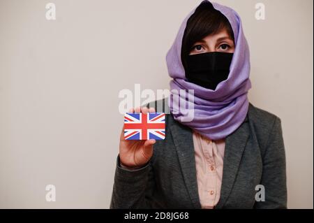 Portrait d'une jeune femme musulmane portant des vêtements formels, protégeant le masque facial et le foulard hijab, tenir la carte drapeau du Royaume-Uni contre un arrière-plan isolé. Banque D'Images