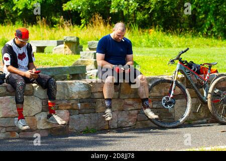 Maryland, Etats-Unis 06/27/2020: Deux hommes reposent sur un mur de pierre dans une forêt et regardent des téléphones. Ils portent des vêtements de cyclisme et ils viennent de terminer Banque D'Images