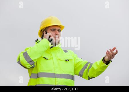 Jeune ingénieur ou travailleur avec des vêtements de travail de protection verte parler sur un téléphone portable devant un ciel lumineux Banque D'Images