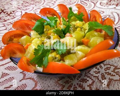 Sur une nappe de dentelle rouge et blanche se trouve un plat bleu rempli de salade de pommes de terre composée de pommes de terre blanches, de tomates, de pois verts, de maïs et de persil avec du mayonnaise Banque D'Images