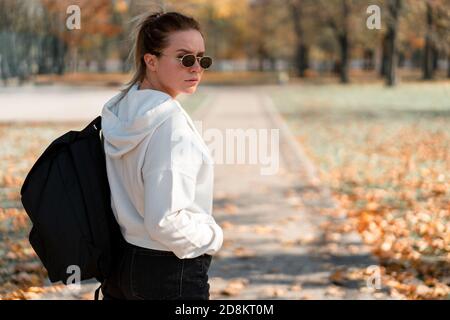 Une belle jeune femme avec une queue de cheval et lunettes de soleil, avec un sac à dos sur ses épaules dans le parc. Photo de l'arrière. Sweat blanc Banque D'Images