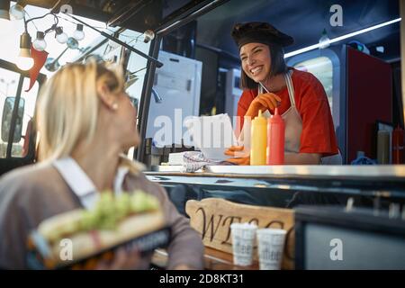 jeune femme caucasienne satisfaite qui s'adresse à un client qui part en sandwich entre les mains et se tourne la tête vers l'employé Banque D'Images