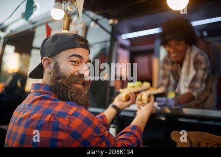 un homme de race blanche satisfait, satisfait, avec un client de barbe qui reçoit des sandwichs d'un employé poli dans le service de restauration rapide, en tournant la tête, en regardant la caméra Banque D'Images