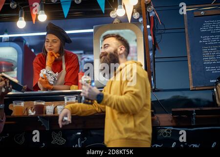 jeune beardy hipster manger un sandwich devant le service de restauration rapide, en socialisant avec une employée féminine Banque D'Images