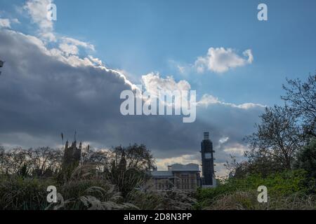 Une vue lointaine de Big Ben en réparation, enveloppée par un échafaudage et prise à travers la verdure, avec ciel bleu et nuages avec des puits de lumière Banque D'Images