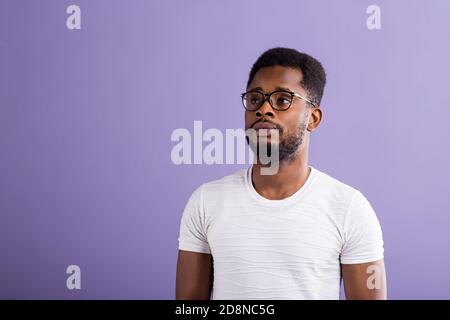 Portrait de l'intelligent calme jeune homme noir barbu à lunettes, vêtu de vêtements décontractés à avec perplexité côté standing against purple studi Banque D'Images