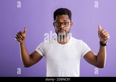 Satisfait homme noir à showing Thumbs up lunettes avec les deux mains, souriant, à côté de blanc portant des t shirt, standing against purple backg Banque D'Images