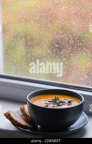 soupe à la crème de citrouille et de carotte avec graines de citrouille et pain grillé. sur le rebord de la fenêtre, avec gouttes de pluie douillettes Banque D'Images