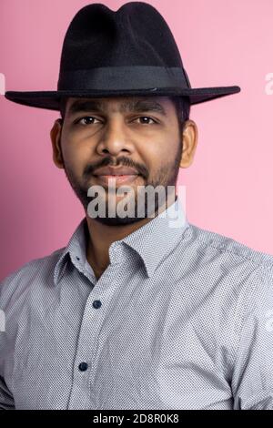 Gros plan de jeune homme indien joyeux et heureux avec barbe de 3 jours, portant une chemise décontractée, un chapeau noir, regardant avec le sourire et pointant sur le côté avec l'index Banque D'Images