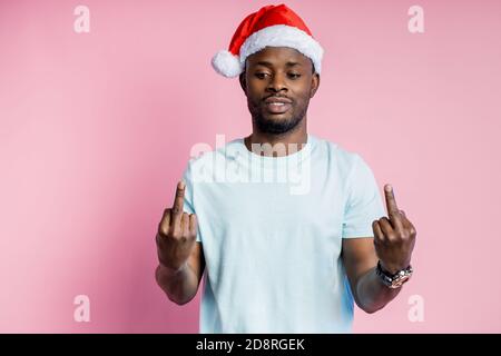 Drôle joyeux afro-américains non rasé jeune étudiant, tout sourire, wearing Santa hat, white t shirt, montrant le majeur des deux mains, isola Banque D'Images