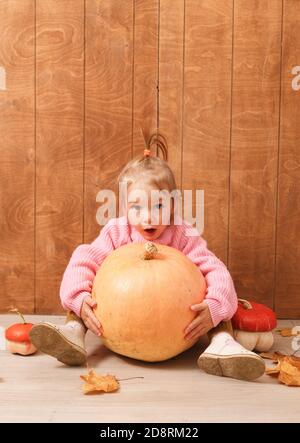 une petite fille mignonne dans un chandail rose enfile un grande citrouille assise sur le sol sur un fond en bois Banque D'Images