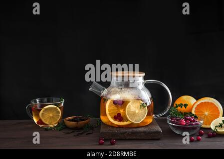 Thé aux agrumes et aux baies, aux canneberges, au citron, aux oranges et au thym, dans une théière en verre sur une table en bois sombre, espace de copie. Thé aux fruits. Saison hiver automne Banque D'Images