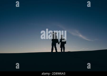 Un jeune couple aventureux tenant les mains, silhoueté contre le coucher du soleil, au sommet d'une colline ou dune. (À Coral Pink Sand Dunes, Utah, États-Unis) Banque D'Images