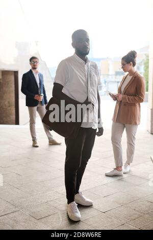 Jeune homme d'affaires africain avec sac à main en cuir debout devant collègues Banque D'Images