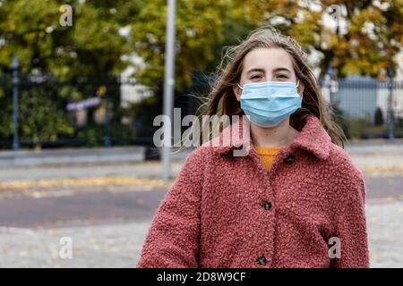 Une fille blonde en manteau d'hiver rose marchant avec confiance dans une rue urbaine portant un masque pour la pandémie du virus Corona. Banque D'Images