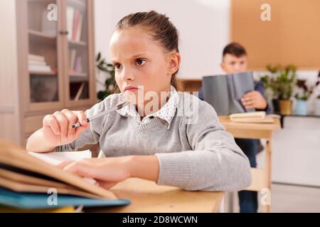 Fille d'école pensive et frognante regardant la tâche dans le livre et penser à la réponse Banque D'Images