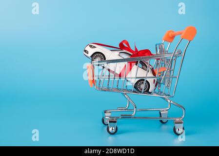 Photo de profil pleine longueur latérale d'une petite voiture miniature jouet avec ruban rouge porté par chariot isolé sur bleu couleur de fond Banque D'Images