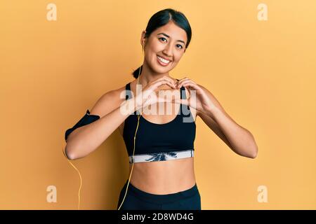 Belle jeune femme de sport asiatique portant des vêtements de sport et des écouteurs souriant dans l'amour montrant le symbole de coeur et la forme avec les mains. Concept romantique. Banque D'Images