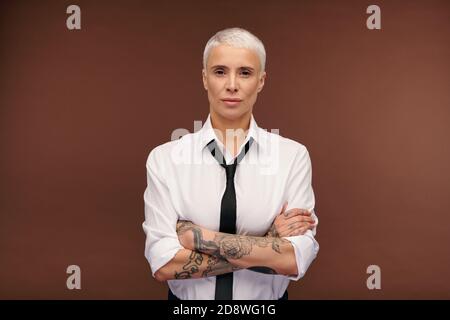 Femme d'affaires blonde sérieuse et confiante croisant les bras avec des tatouages poitrine Banque D'Images