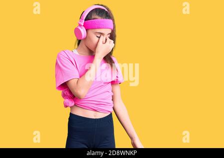 Adorable enfant hispanique fille portant des vêtements de gym et utilisant des écouteurs fatigués frottant nez et les yeux sensation de fatigue et de maux de tête. Stress et frustration con Banque D'Images